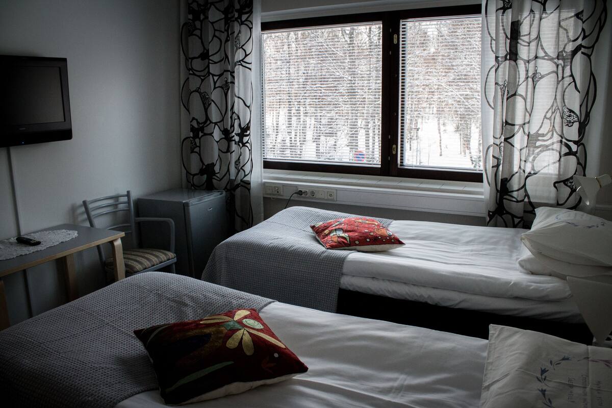 Quarto de hotel com duas camas, uma escrivaninha com cadeira, televisão e uma janela com cortinas.