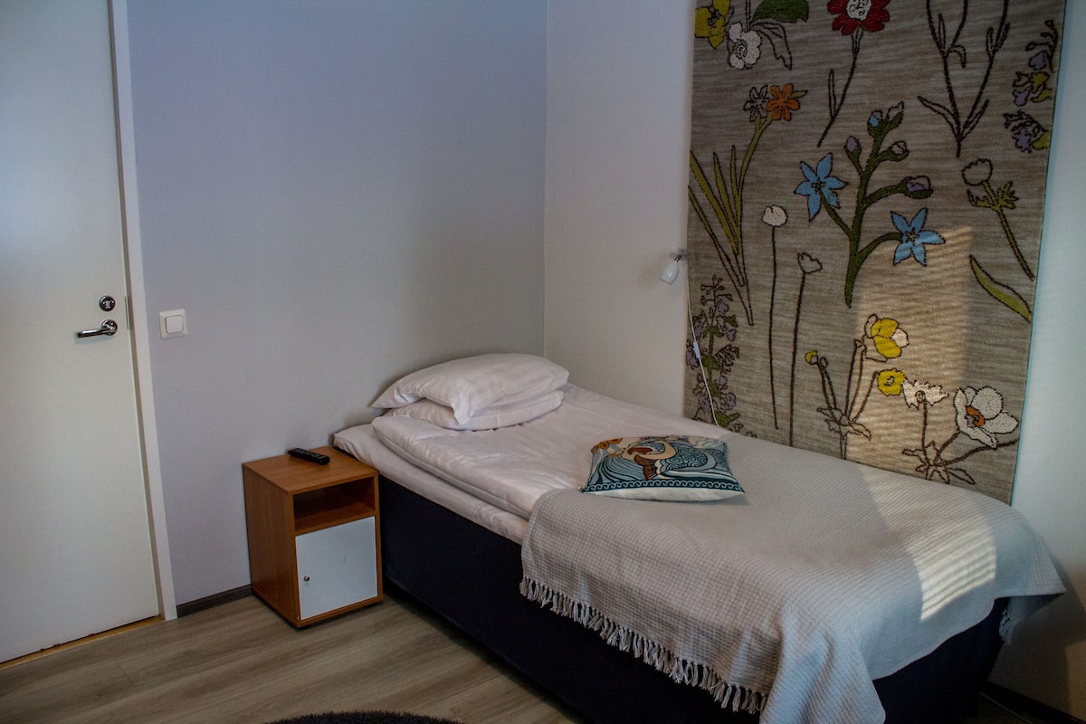 Um quarto pequeno com uma cama, uma mesinha de cabeceira, uma tapeçaria na parede e uma porta.