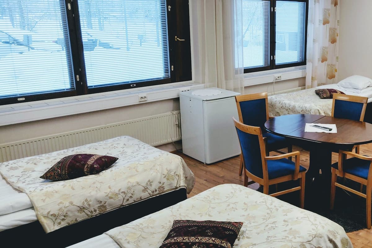 Quarto de estudante com duas camas individuais, uma mesa pequena com cadeiras, um radiador e uma janela com persianas.