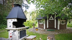A barbecue grill with a chimney, a small wooden structure, and a picnic table in a garden setting. - Hotelli Vuolake (Laukaa)