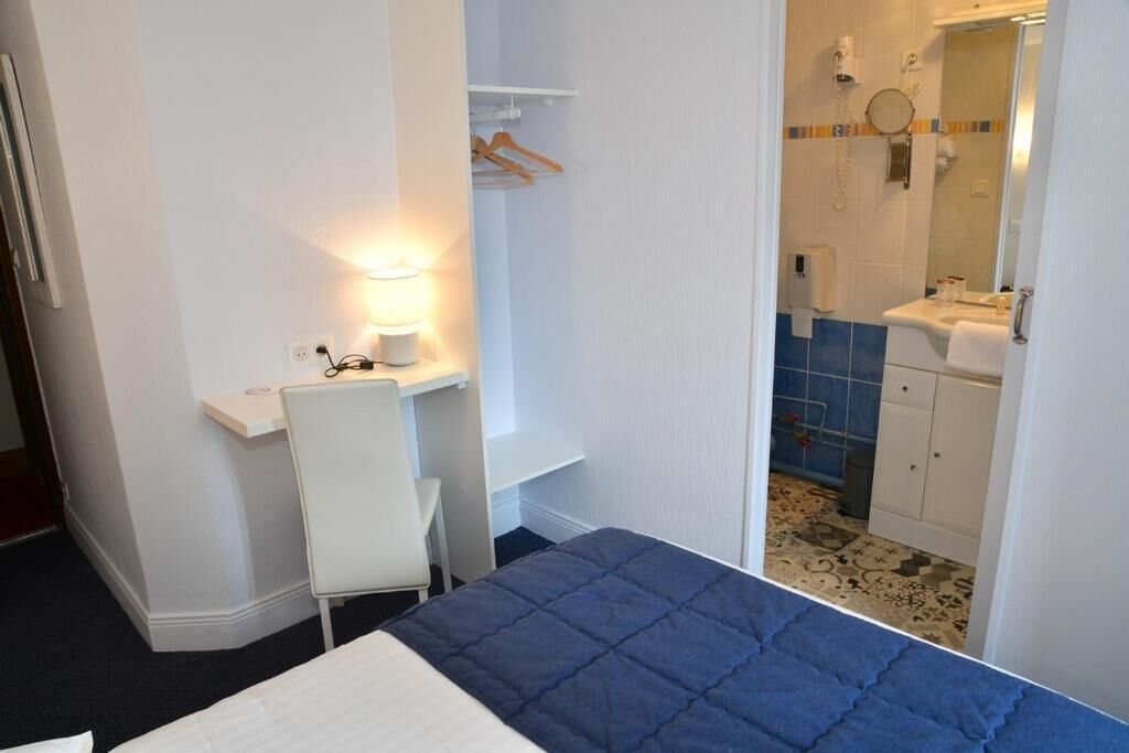 A hotel room with a bed, a desk with a lamp, a chair, a bathroom with a sink and mirror, and a shower.