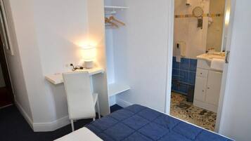 A hotel room with a bed, a desk with a lamp, a chair, a bathroom with a sink and mirror, and a shower.