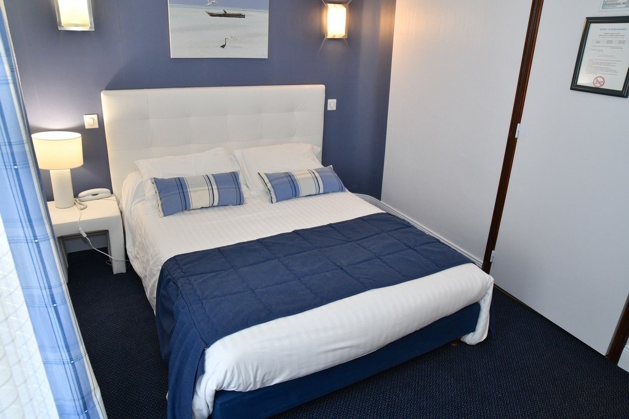 Una cama bien tendida con cabecera blanca y ropa de cama azul, una lámpara de noche y un certificado enmarcado en la pared.