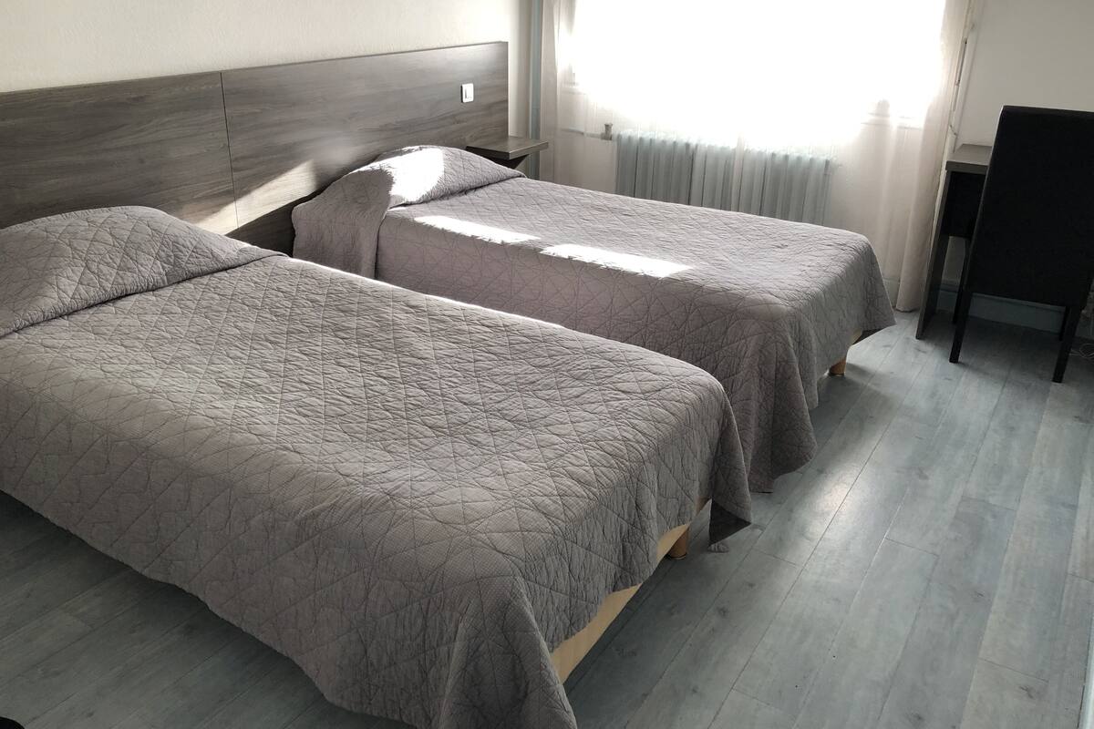 A room with two single beds, a wooden headboard, a window with sheer curtains, and a chair.