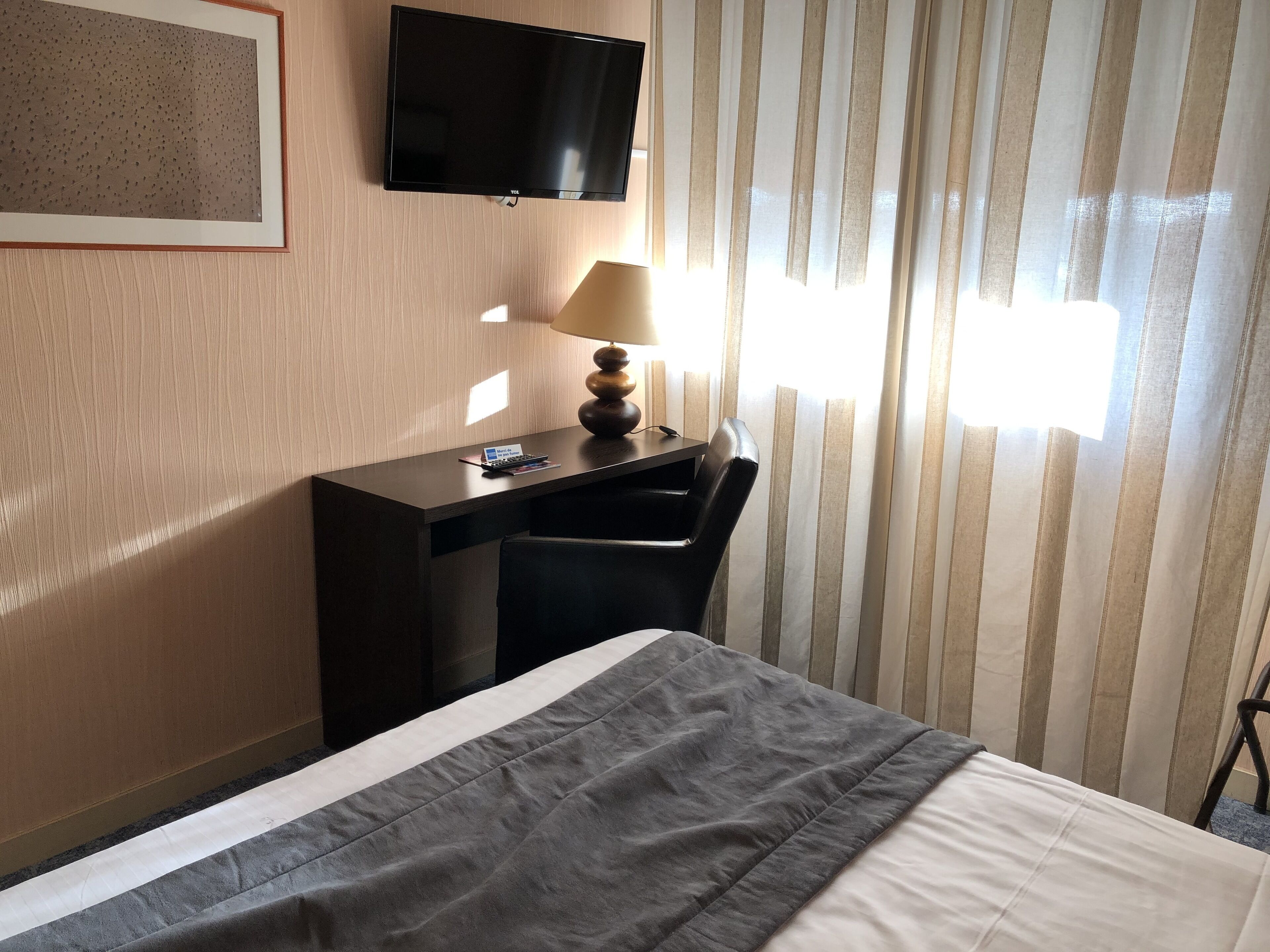 A hotel room with a bed, a desk with a lamp, a chair, a TV mounted on the wall, and a window with curtains.
