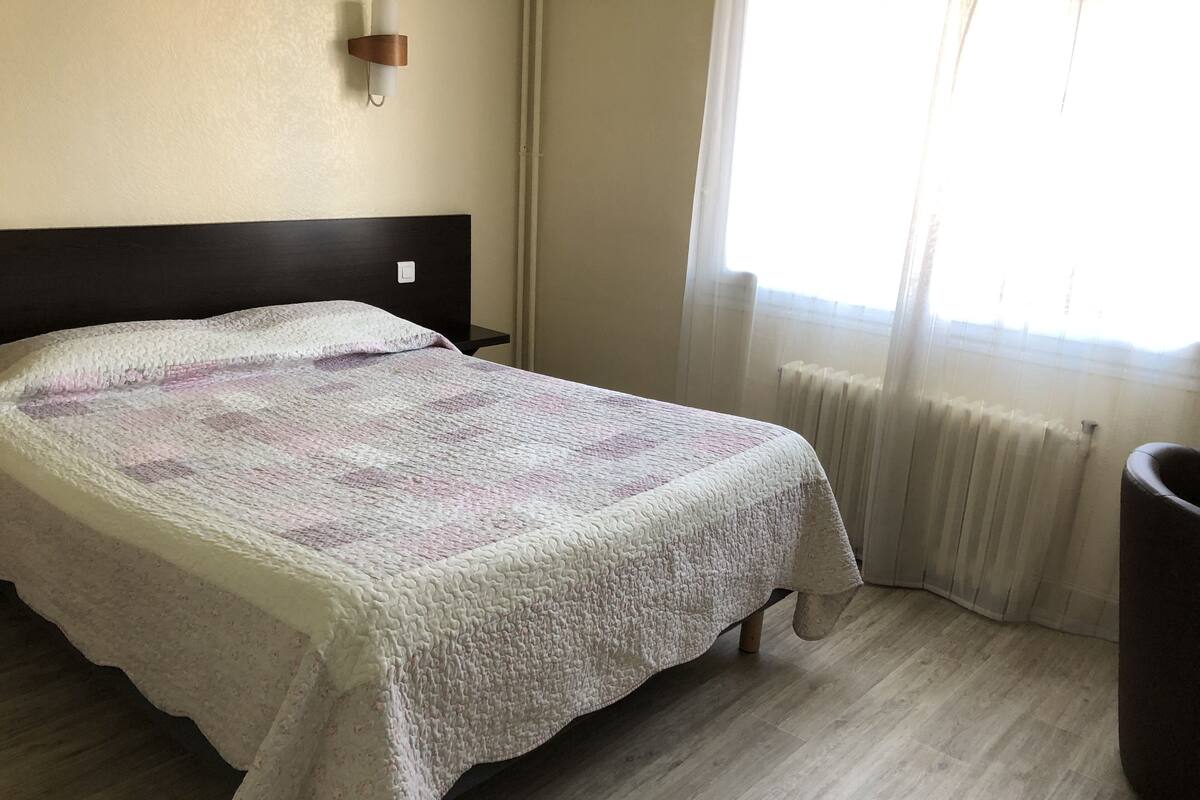 A neatly made bed with a quilt, a bedside table, a wall-mounted lamp, and a radiator.