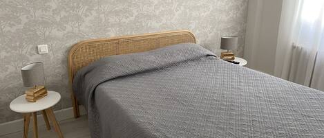 Une chambre à coucher avec une tête de lit en osier, une couvre-lit matelassée grise, une table de chevet avec une lampe et un mur orné d’un motif d’arbre.