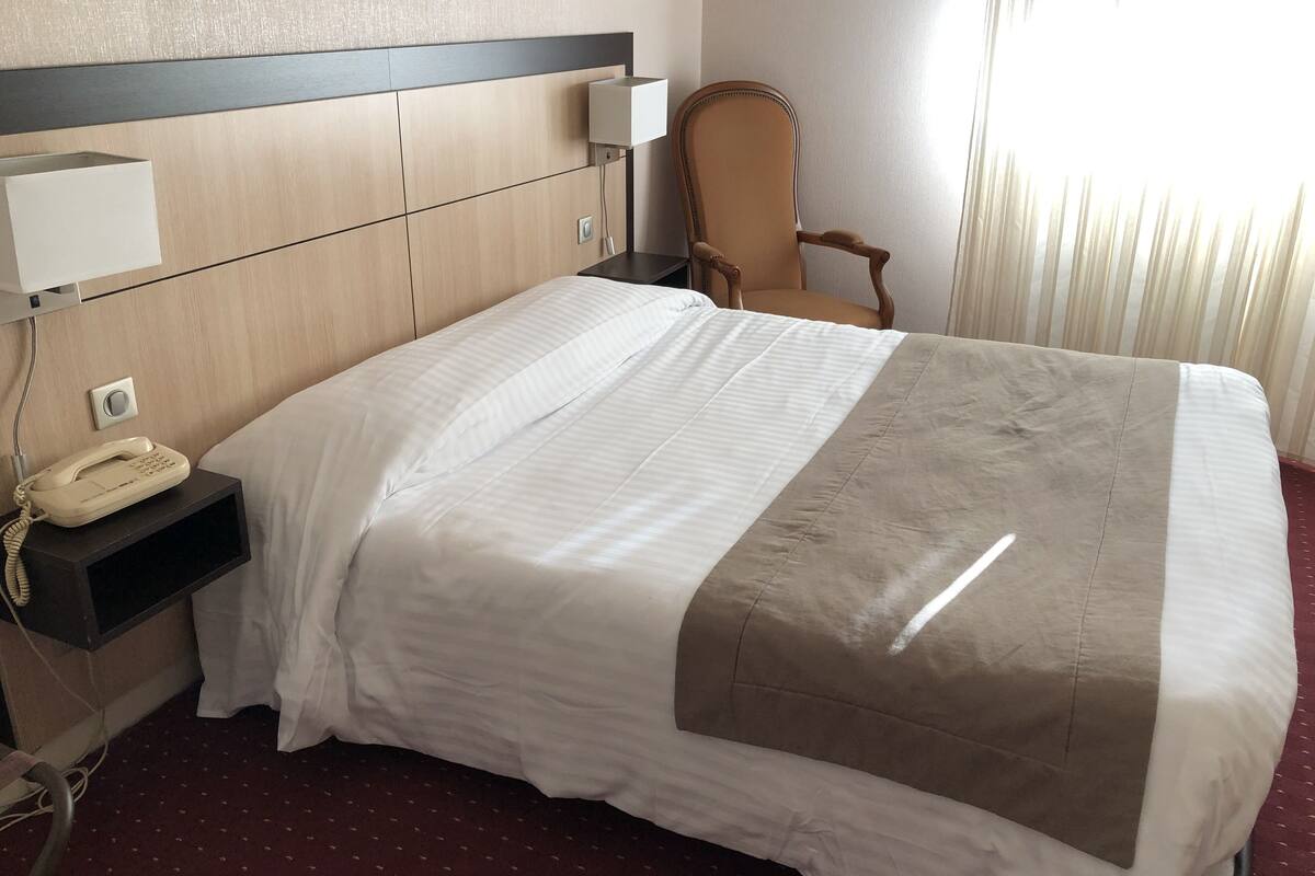 A hotel room with a bed, a telephone, a chair, and a window with curtains.