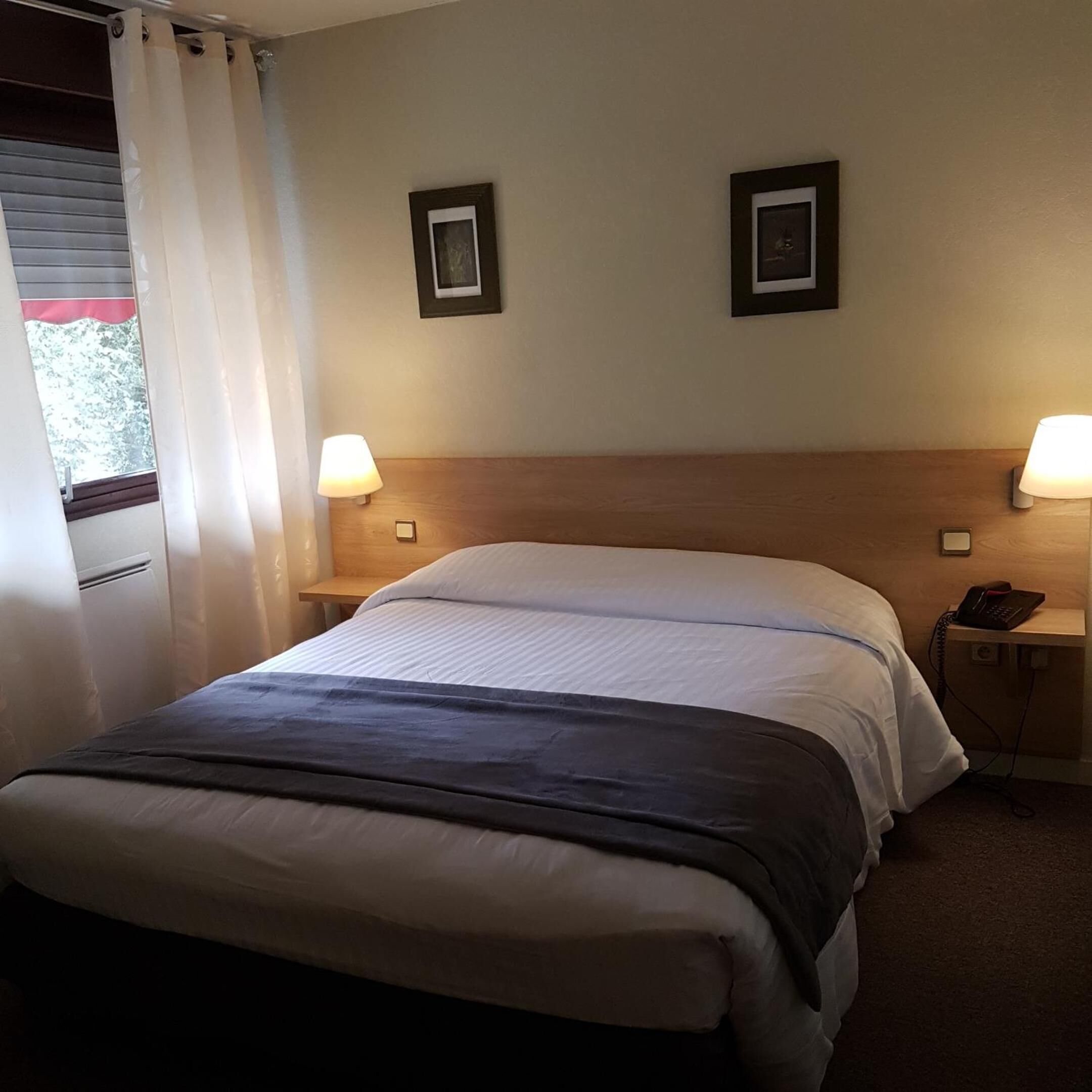 A hotel room with a bed, two bedside lamps, a nightstand with a phone, and two framed pictures on the wall.