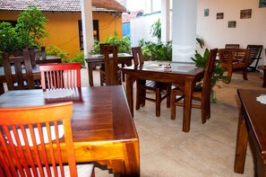 Comedor con mesas y sillas de madera, rodeado de plantas y una pared blanca decorada con obras de arte.