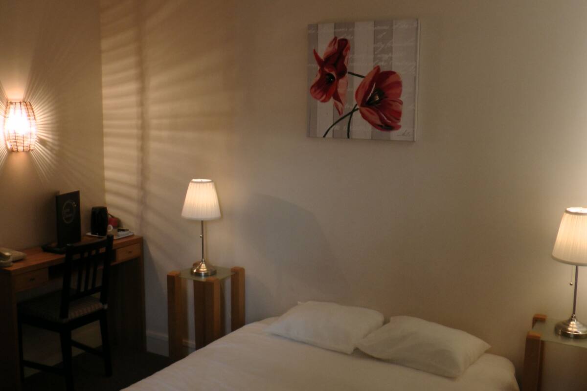 A hotel room with a bed, two bedside lamps, a desk with a chair, a telephone, and a painting on the wall.