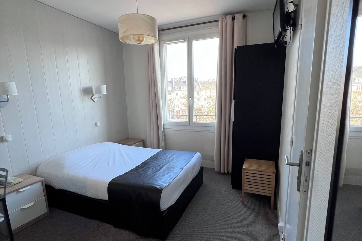 A hotel room with a large bed, a bedside table, a lamp, a window with curtains, and a door.
