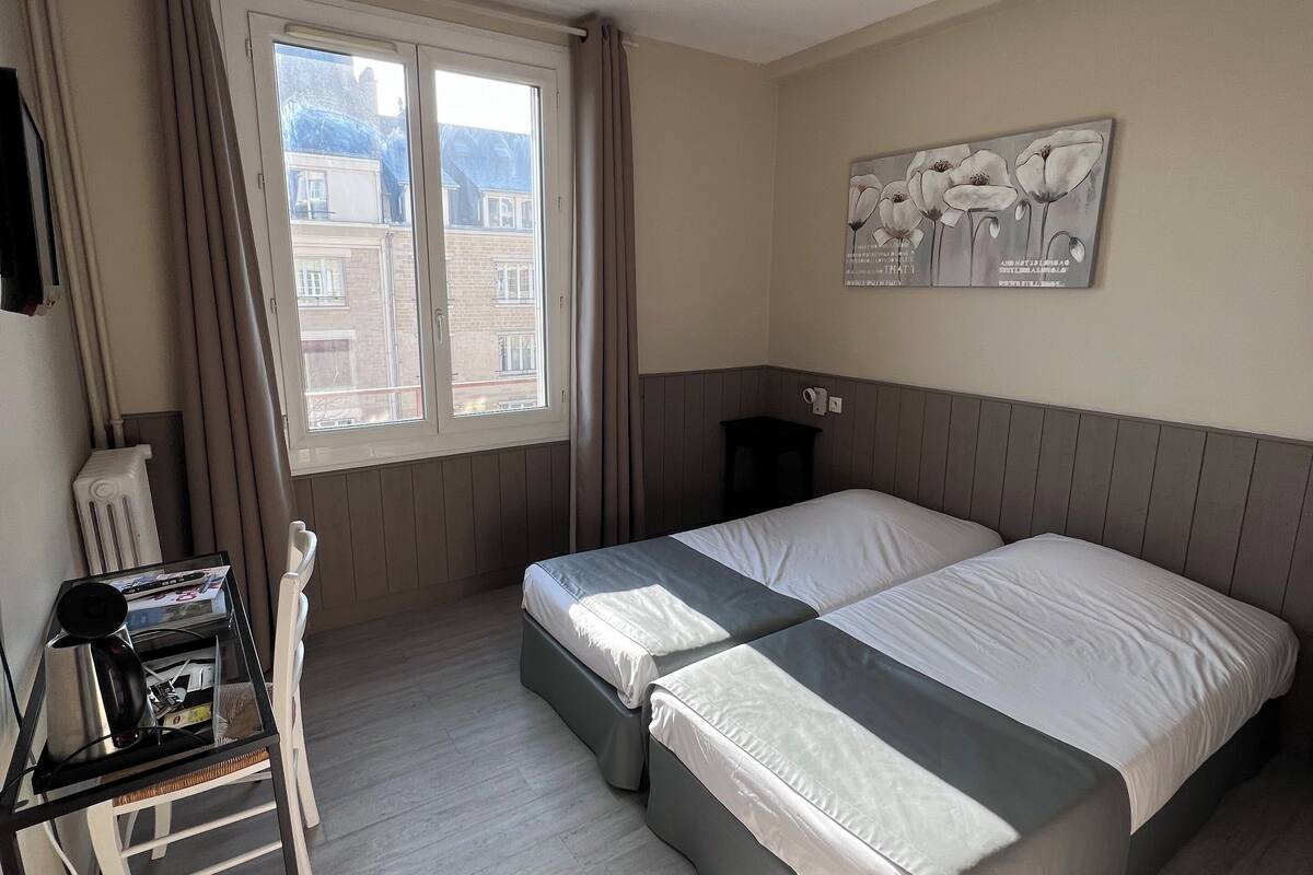 A hotel room with two beds, a desk with a kettle, and a window with curtains.