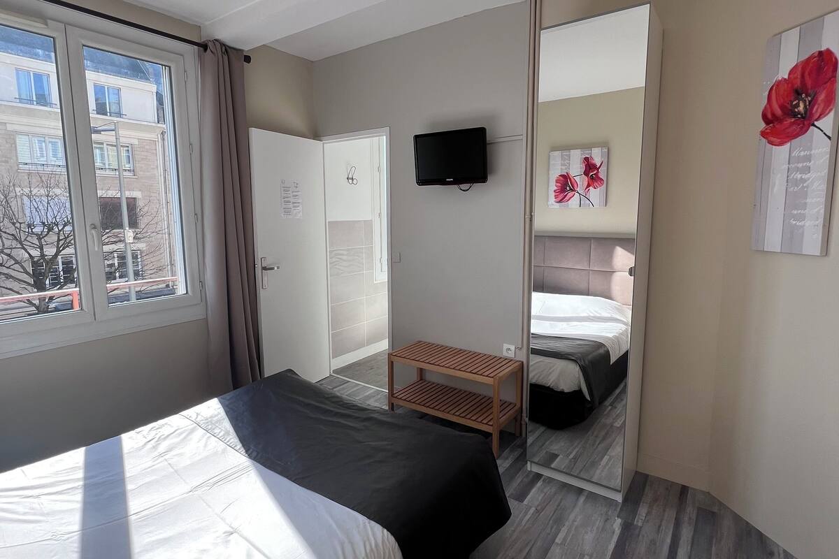 A hotel room with a bed, a TV mounted on the wall, a wooden side table, and a mirror reflecting the room.