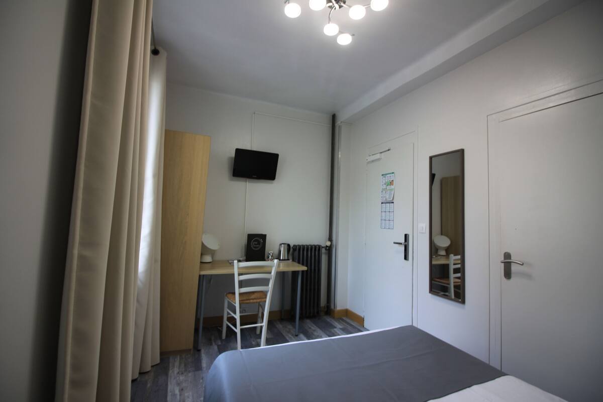 A bedroom with a bed, a desk with a chair, a television, a radiator, and a door with a mirror.