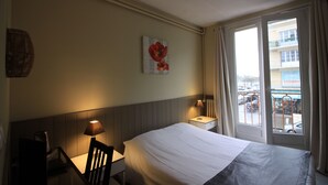 A hotel room with a bed, a desk, a lamp, a window with curtains, and a wall with a flower painting. - Hôtel de L'Univers (Caen)
