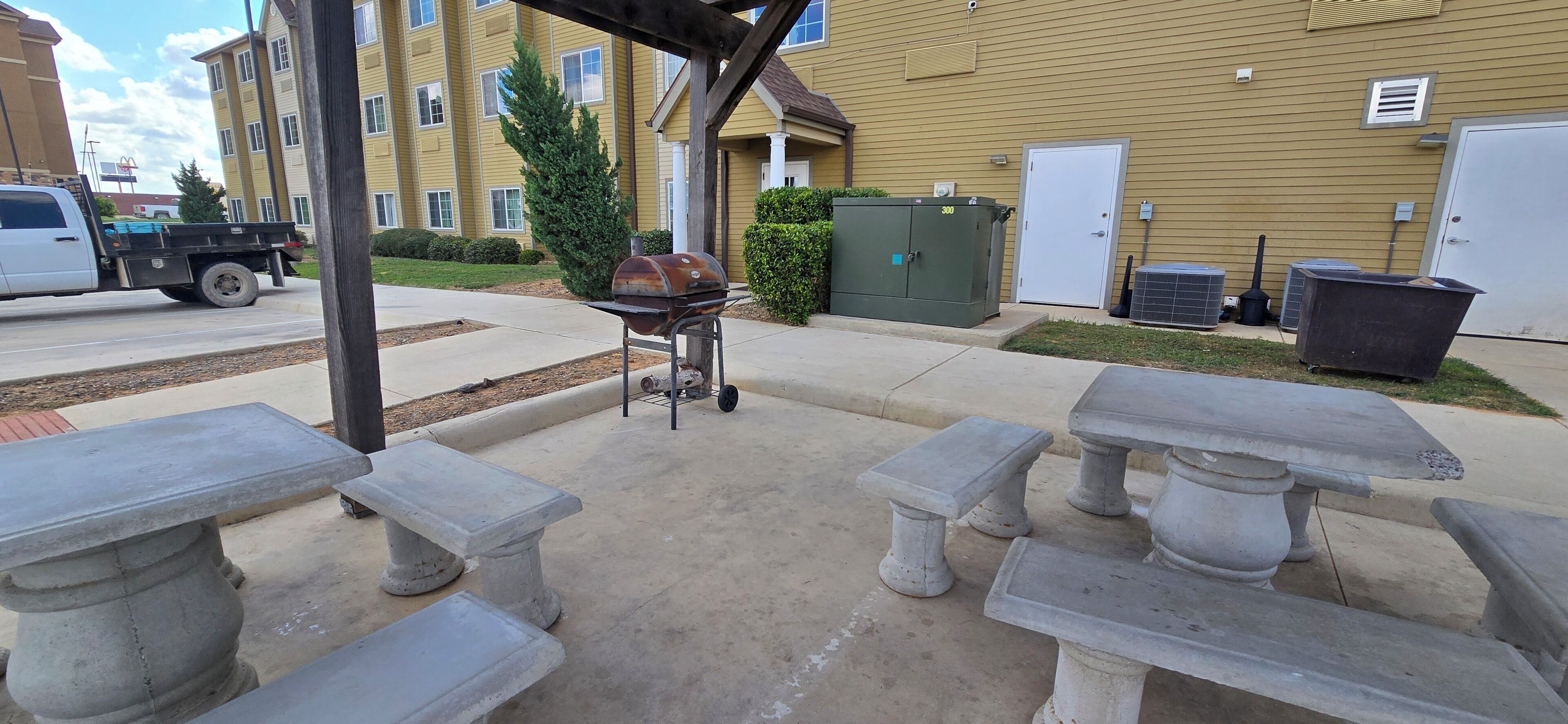 An outdoor patio with concrete tables and benches, a barbecue grill, and a large green utility box.