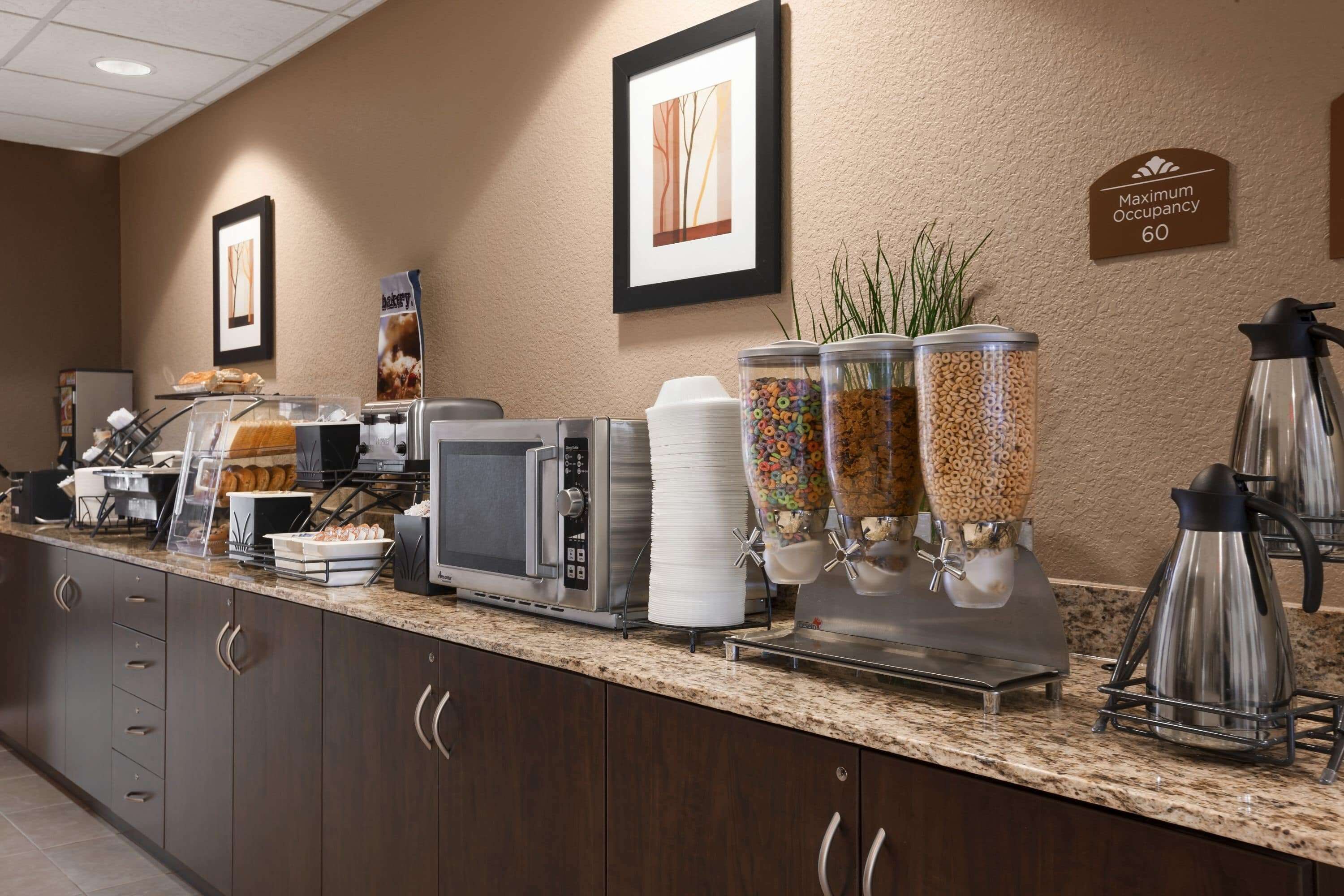 A hotel breakfast area with a coffee maker, cereal dispensers, and a sign indicating maximum occupancy of 60.