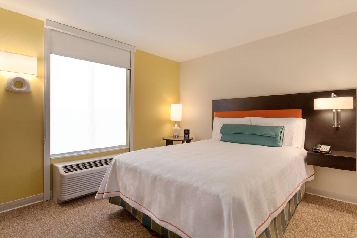 Habitación de hotel con cama, lámparas de noche, calefactor y ventana con persianas.