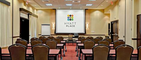 Un salón de conferencias con una pantalla grande mostrando el logotipo de Hyatt Place, filas de mesas y sillas, y piso alfombrado.