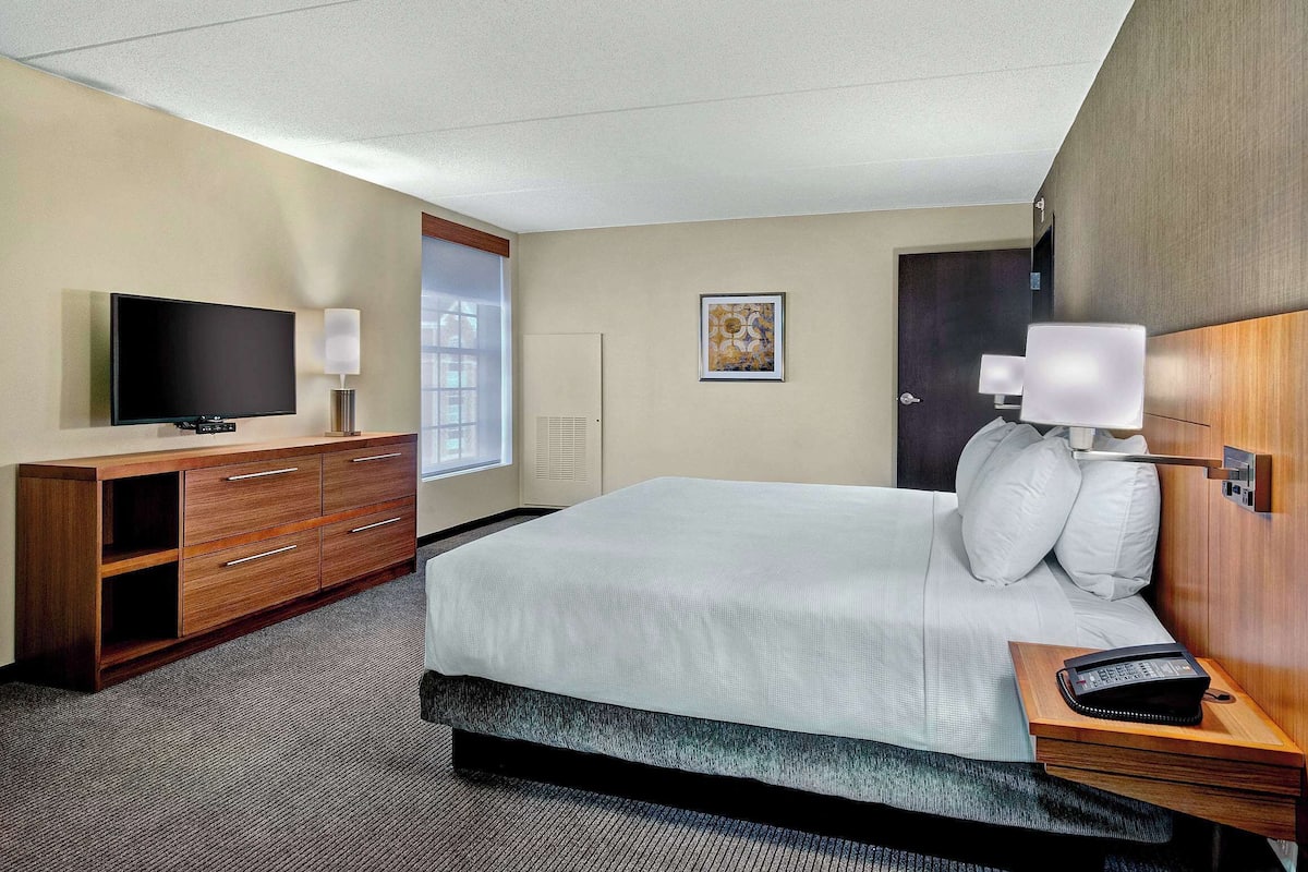 A hotel room with a large bed, a TV, a desk with drawers, a lamp, and a bedside table with a phone.