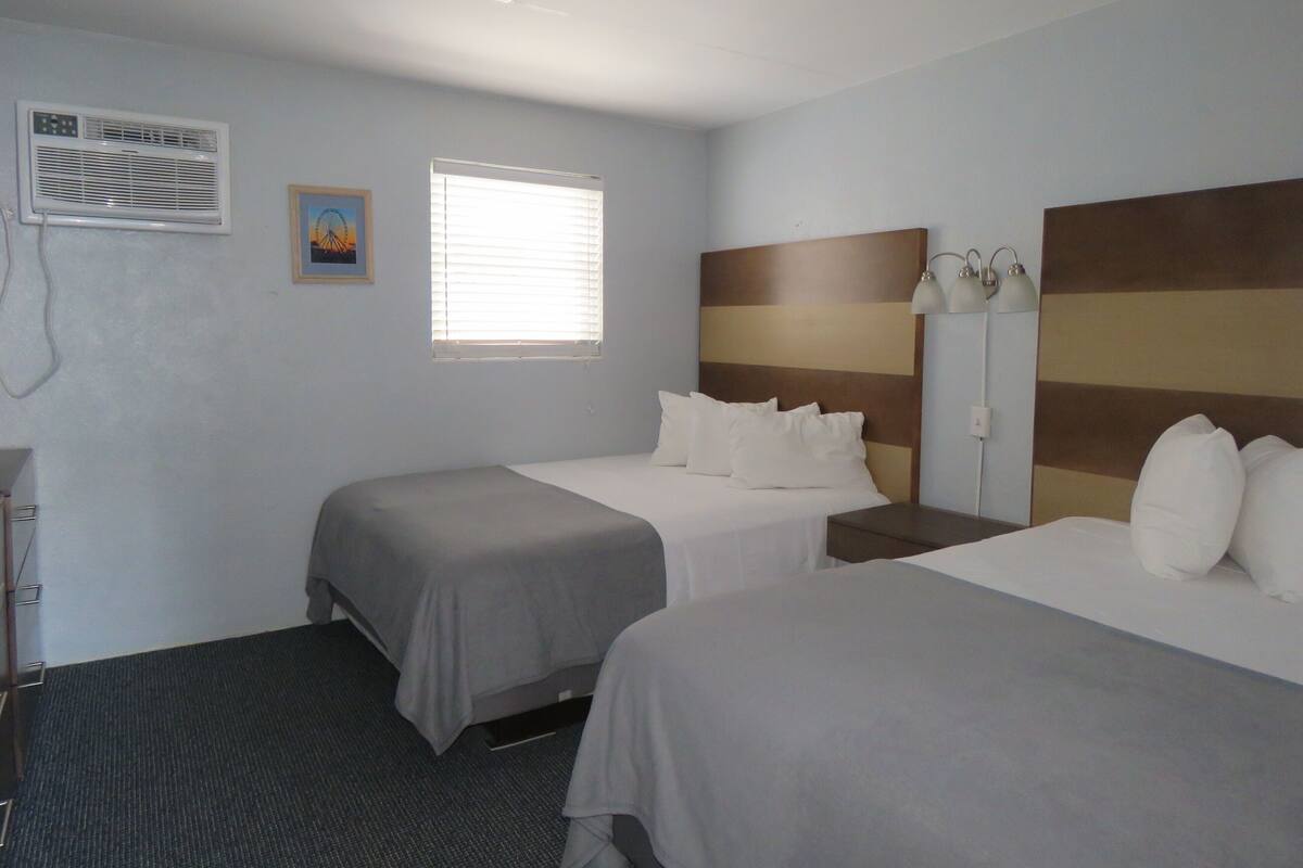 A hotel room with two beds, a window with blinds, an air conditioning unit, and a small painting on the wall.