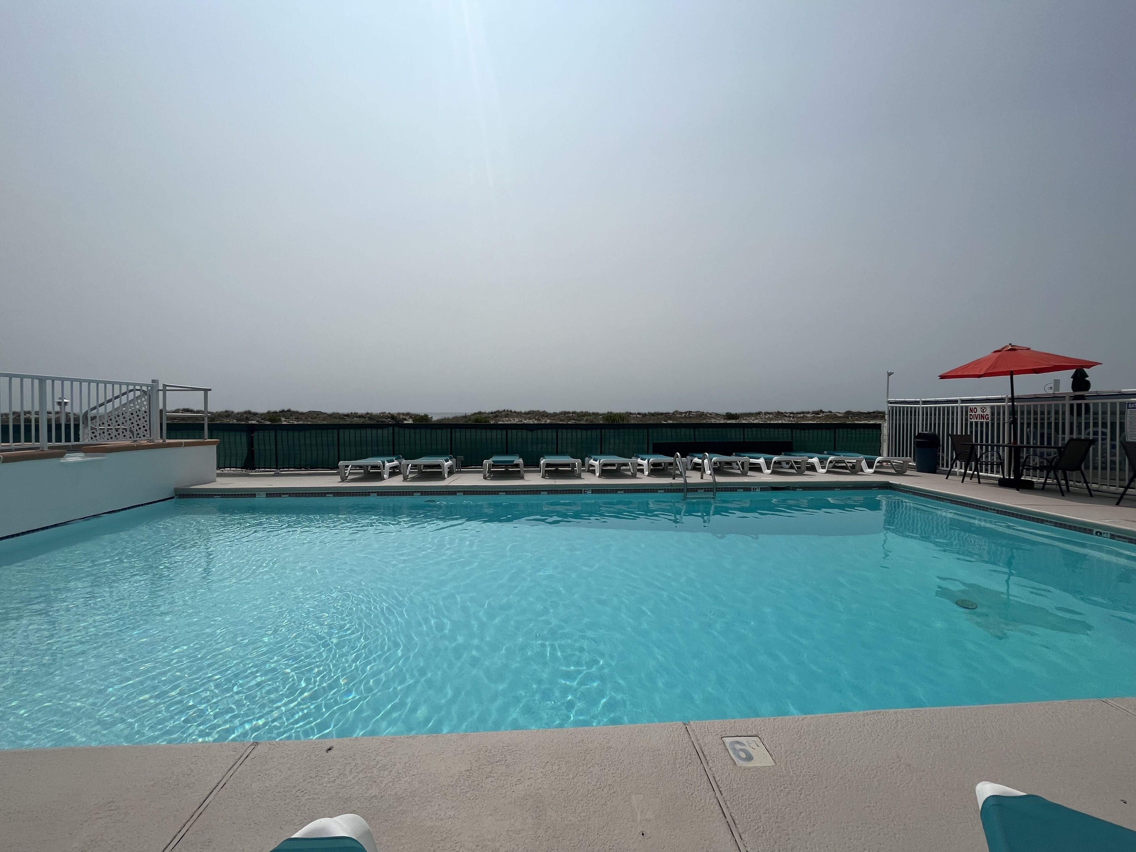 Une piscine aux eaux cristallines, bordée de chaises longues et agrémentée d’un parasol rouge, le tout entouré d  ’une clôture blanche.