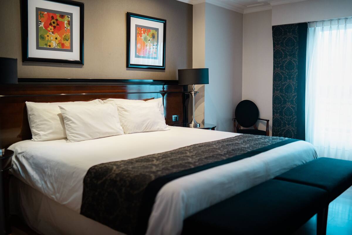 A neatly made bed with white linens and a dark throw blanket, a bedside table with a lamp, and two framed artworks on the wall.