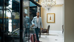 Un hombre con traje azul claro y corbata gris sale del vestíbulo de un hotel con una maleta roja.