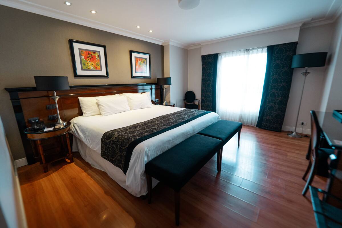 A hotel room with a large bed, a bench, a desk, and two framed pictures on the wall.