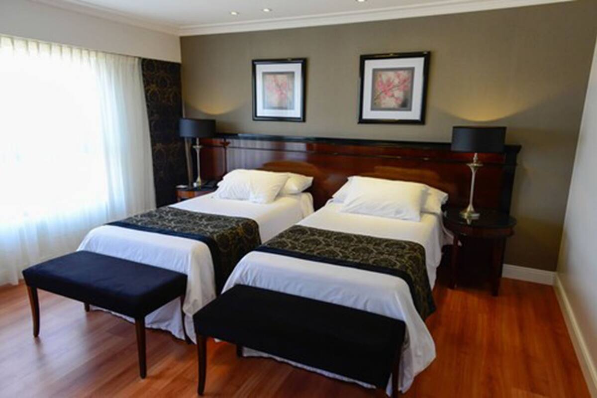 A hotel room with two beds, a bench, a nightstand, and a lamp.