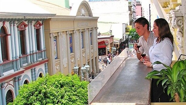 A balcony with a view of a building with classical architecture, including columns and a balcony railing. - Hotel El Maragato (San José)