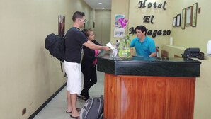 A hotel reception desk with a wooden counter, a man in a blue shirt behind it, and a woman in a black top and white shorts standing in front. - Hotel El Maragato (San José)