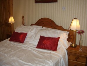 Una cama bien hecha con sábanas blancas y dos almohadas rojas, flanqueada por mesitas de noche de madera con lámparas y un florero con una flor.