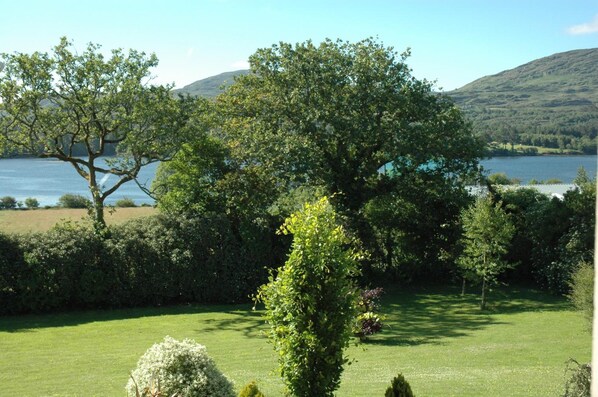 Un jardín exuberante con diversos árboles y arbustos, un césped cuidado y una vista de un lago y colinas al fondo.