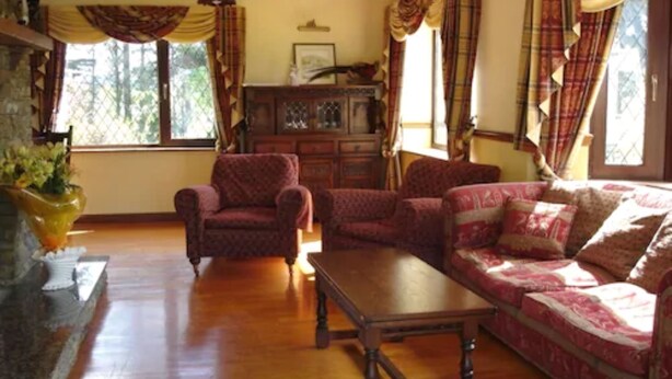 A cozy living room with a wooden floor, a sofa, a chair, and a wooden coffee table. There is a fireplace with a vase of flowers, and curtains on the windows. - Abbey Court (Kenmare)