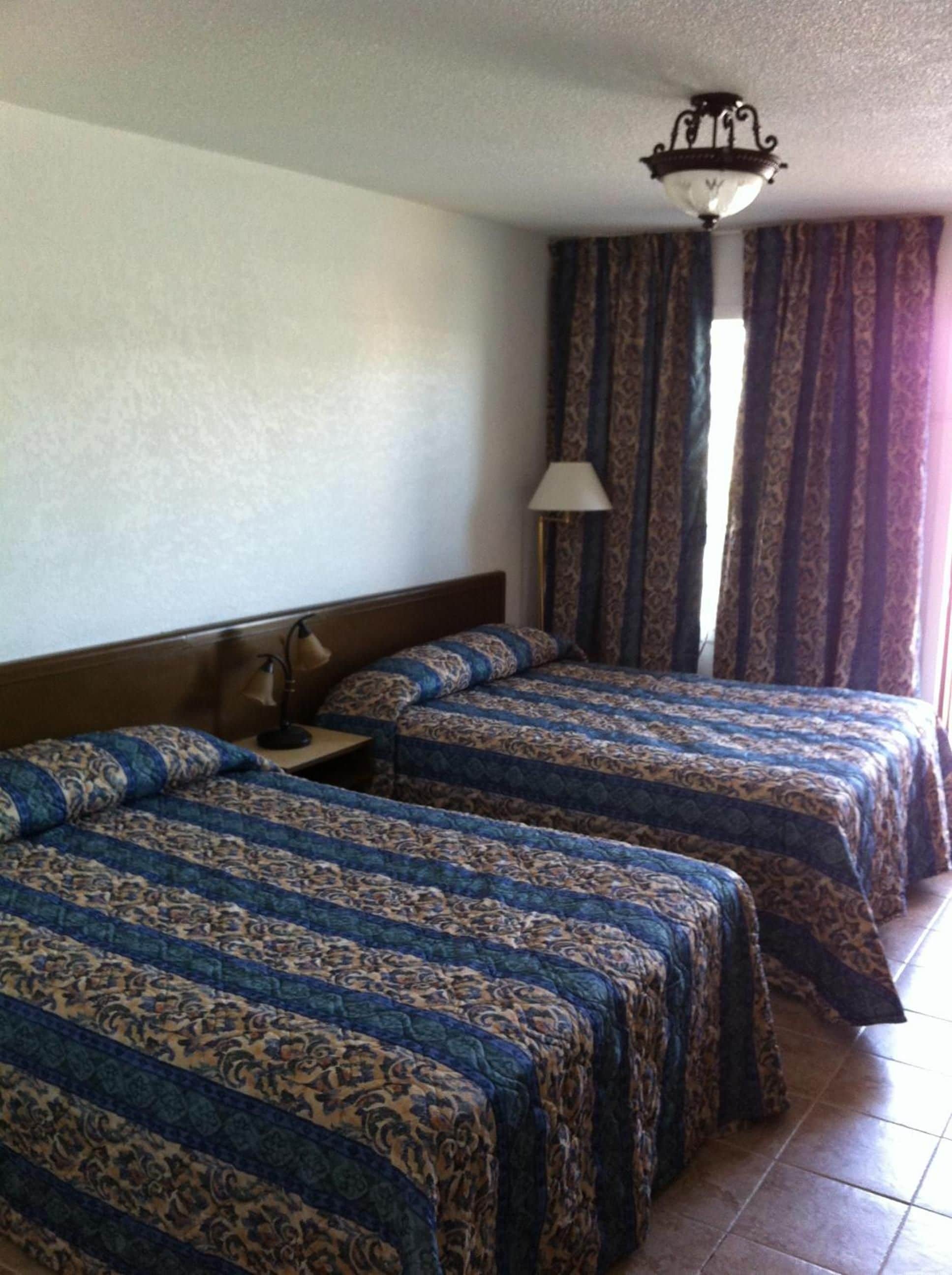Une chambre d’hôtel avec trois lits, une table de chevet, une lampe et une fenêtre avec des rideaux.
