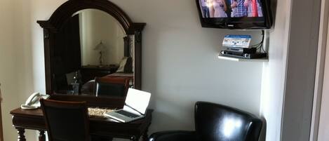 A hotel room with a desk, laptop, chair, TV, and a mirror.