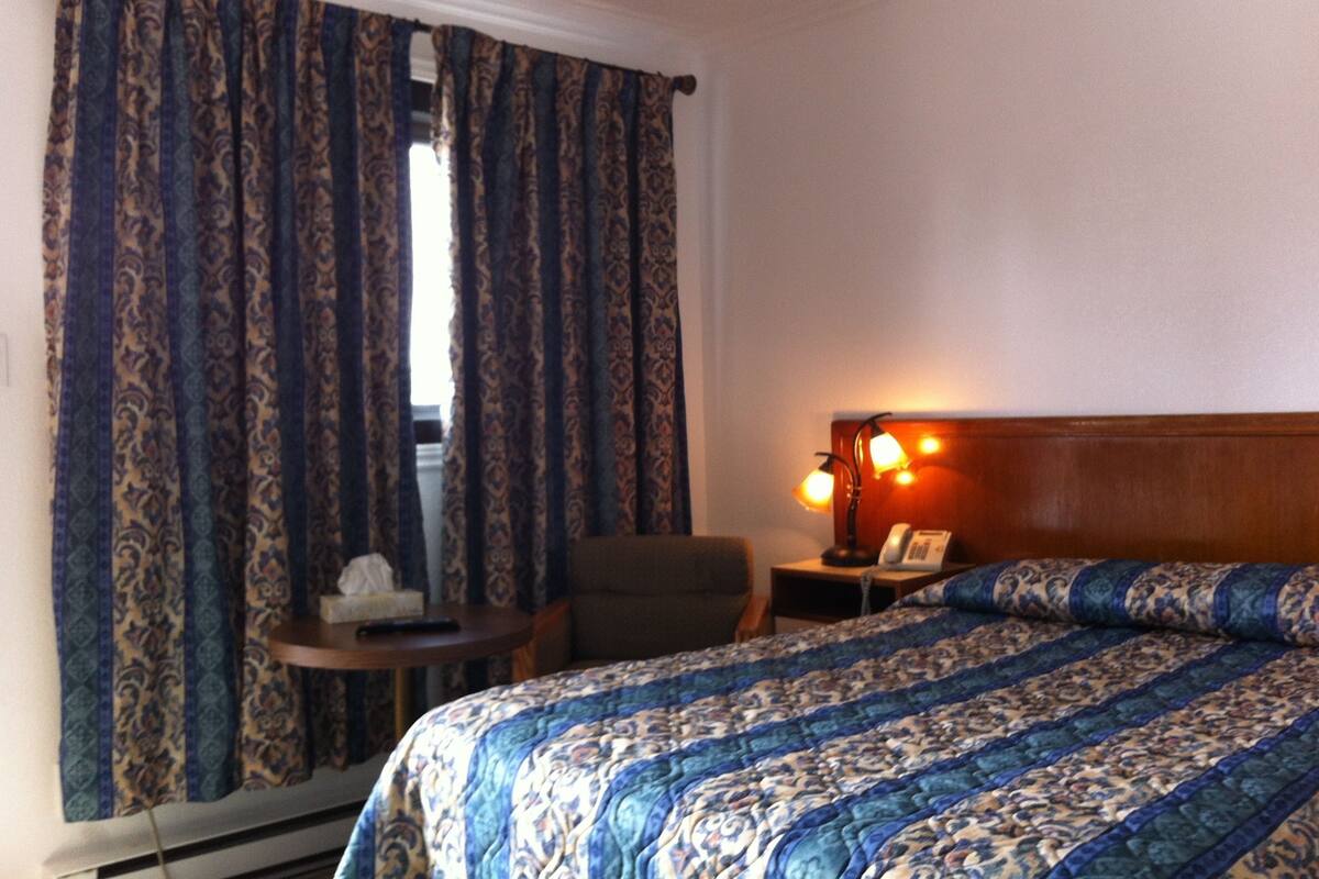A hotel room with a bed, a chair, a small table, and a nightstand with a lamp.