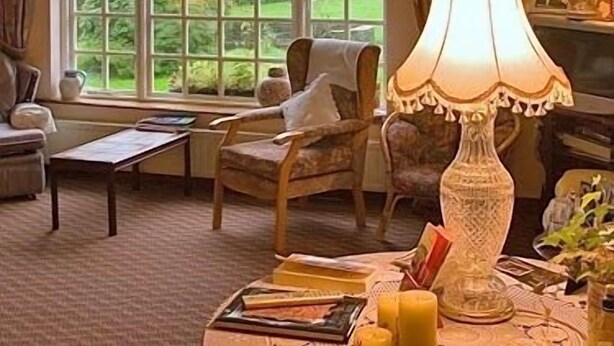 A cozy living room with a large window, a wooden chair, a table with books and a candle, and a decorative lamp. - Salmon Leap Farm B&B (Killarney)