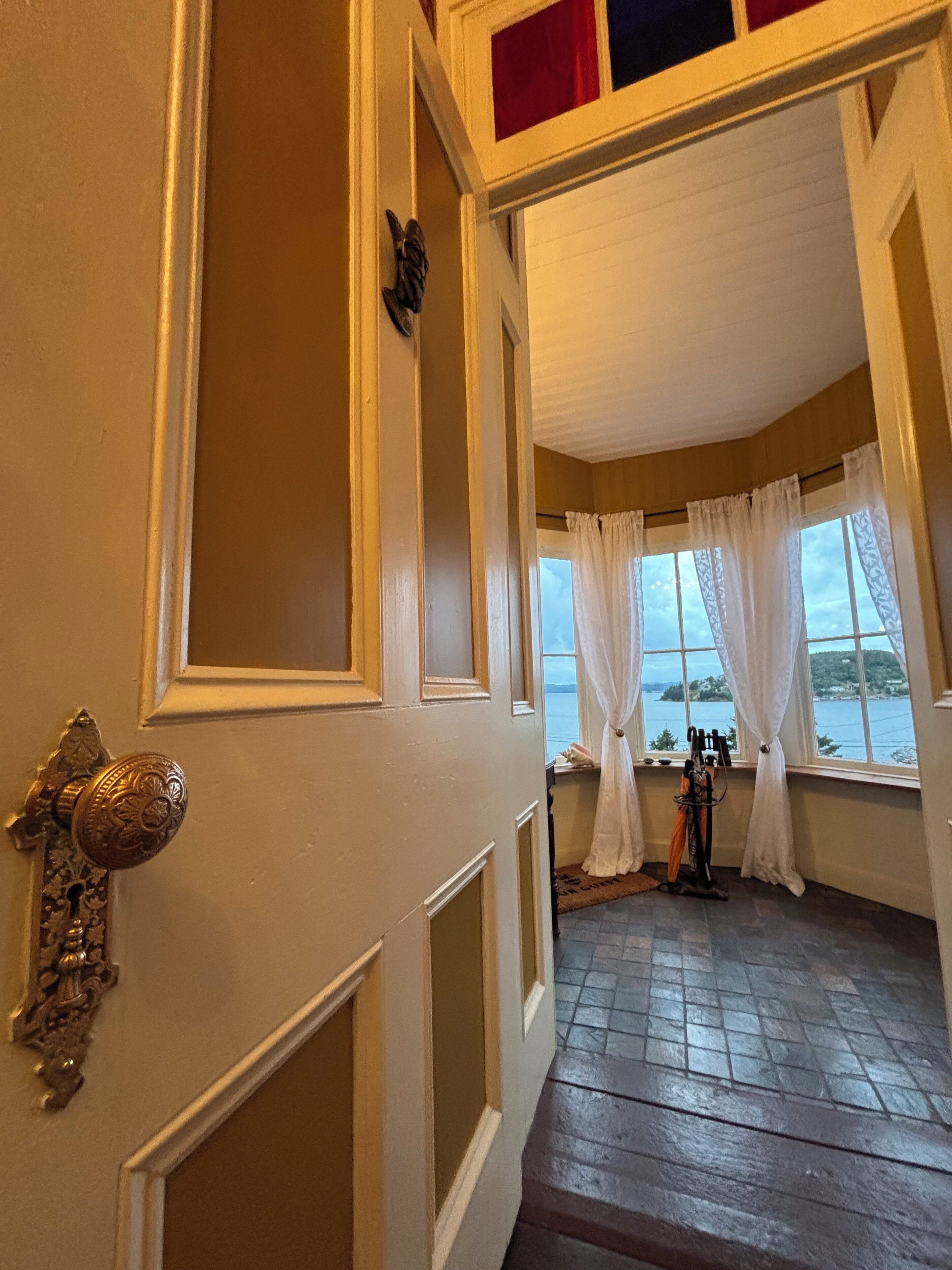 Una habitación con piso de madera, una puerta con pomo decorativo y vista a un cuerpo de agua a través de una ventana con cortinas blancas.