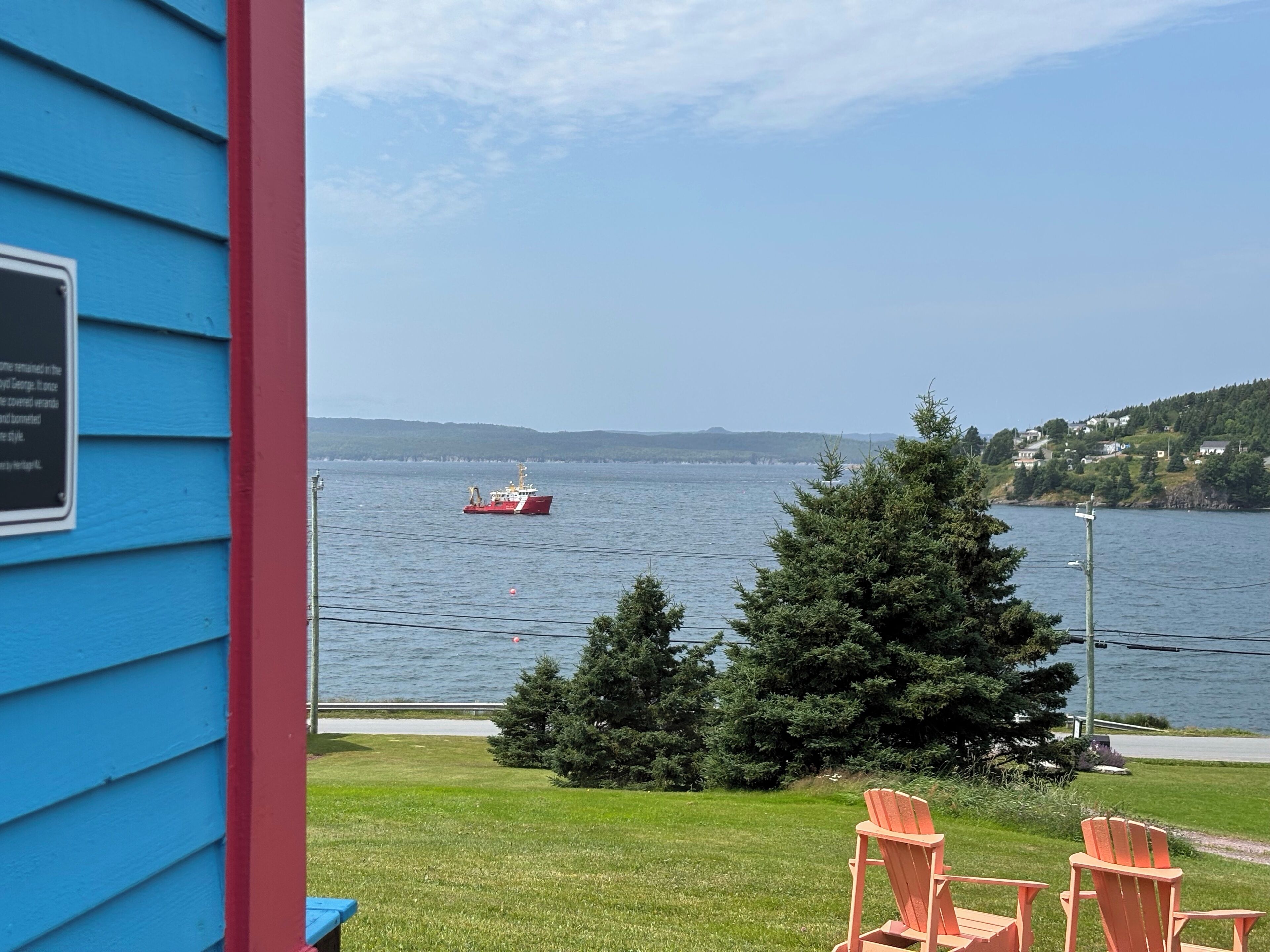 Une vue depuis un porche, avec une maison bleue, un bateau rouge sur l’eau et un paysage verdoyant, où l’on aperçoit des arbres et des maisons au loin.
