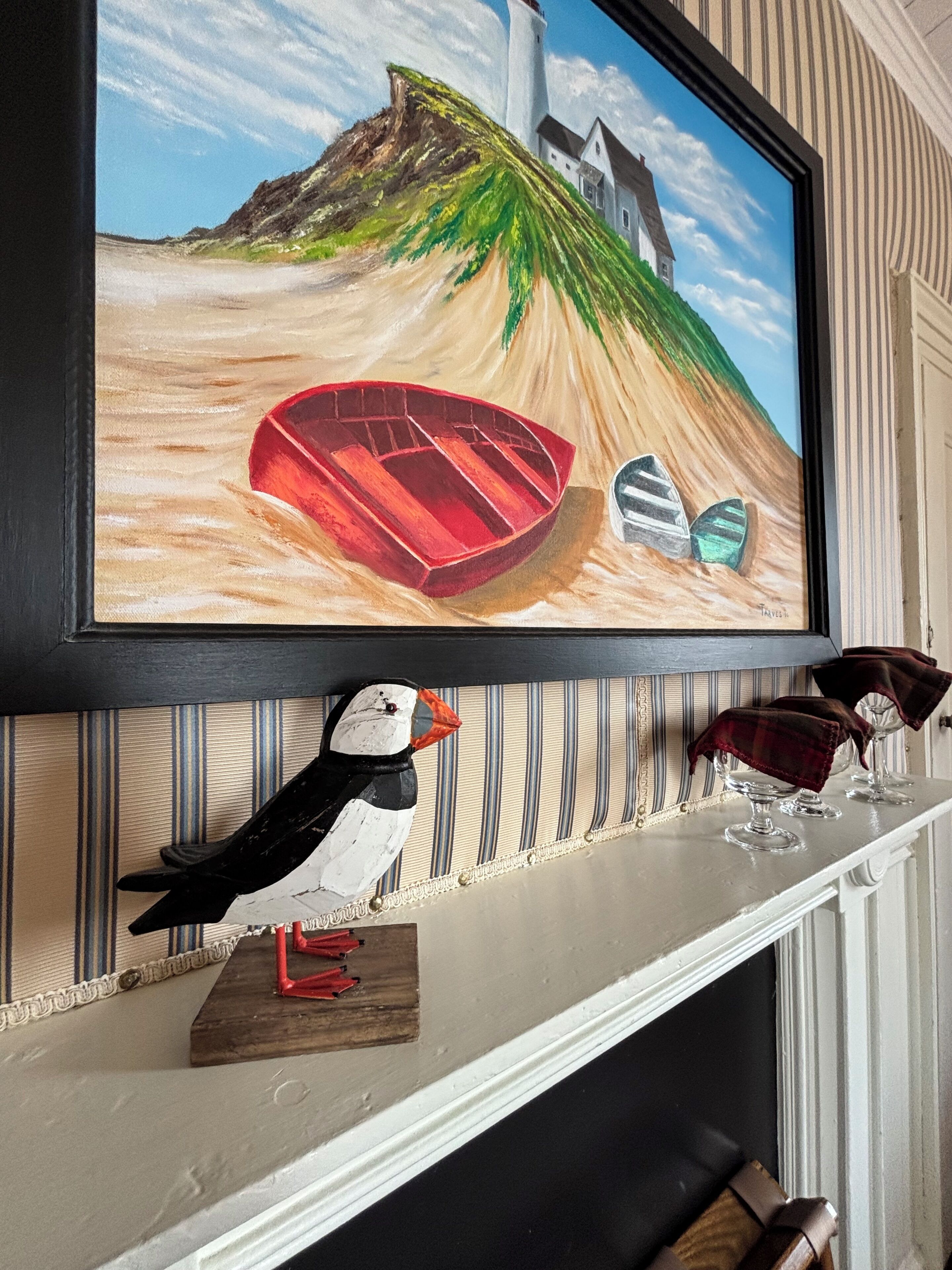 A television mounted on a wall displaying a painting of boats on a beach, with a pufferfish and a small wooden piece on the shelf below.