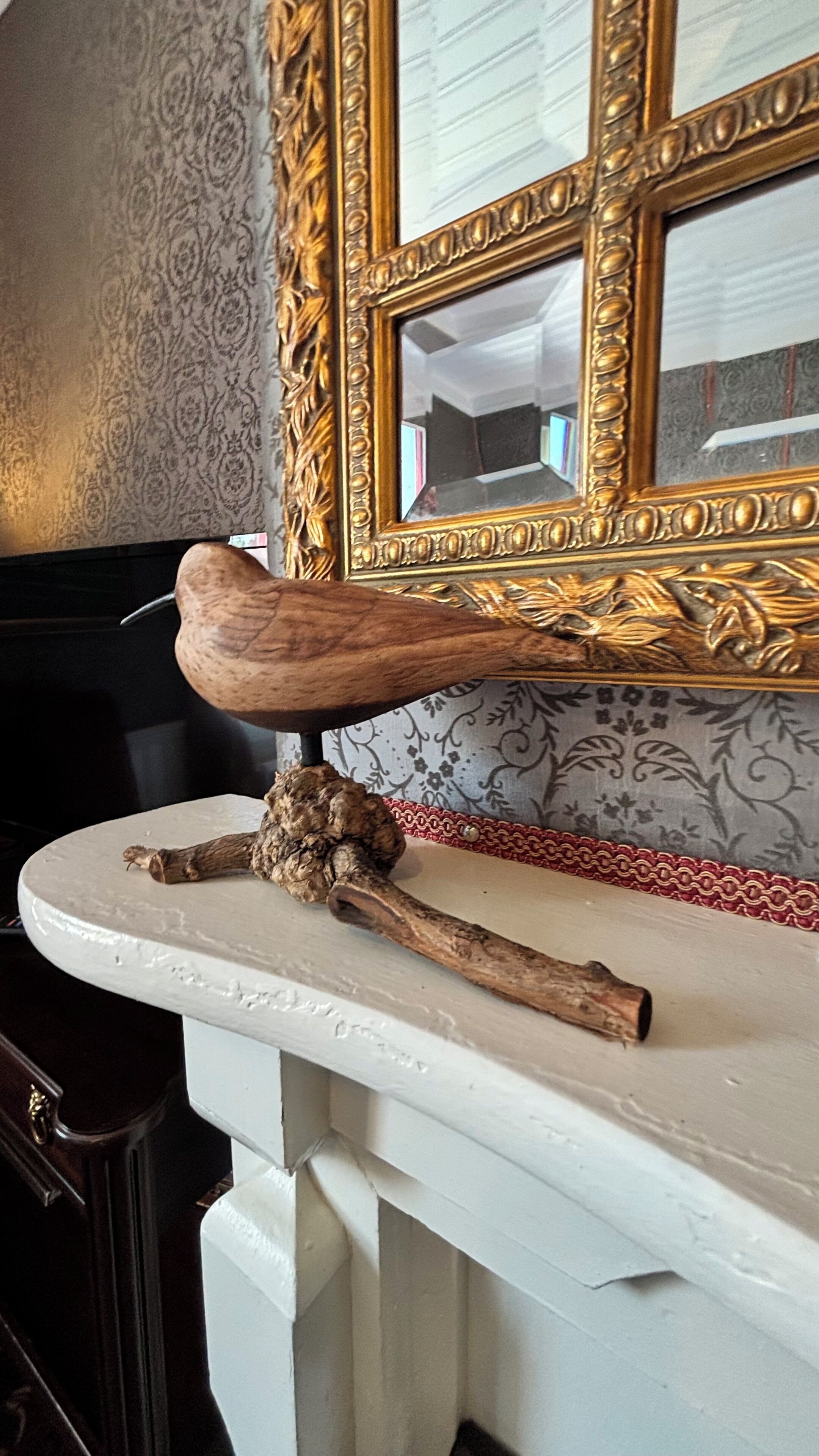 A wooden object on a white surface, a mirror with a gold frame, and patterned wallpaper.