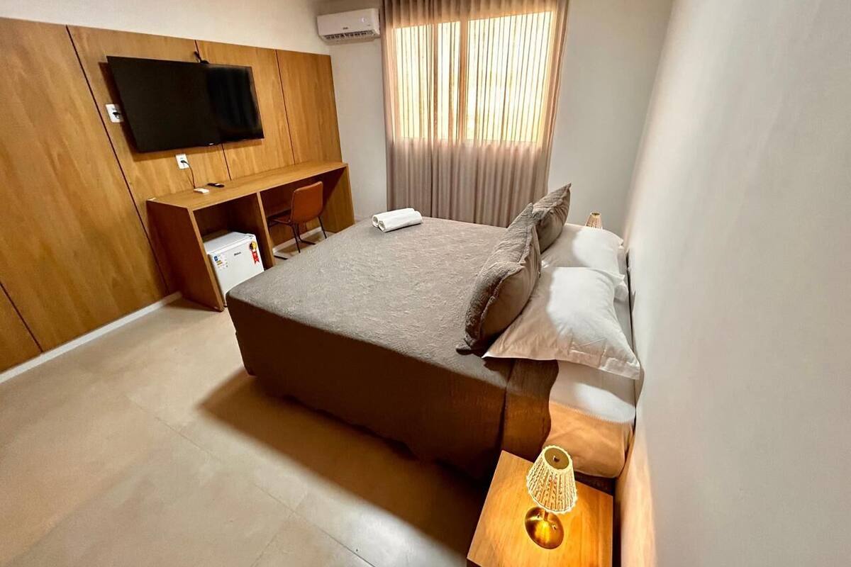 Un dormitorio con cama, televisor montado en la pared, escritorio con silla y ventana con cortinas.