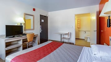 A hotel room with a bed, a desk with a computer, a chair, a small table, and a bathroom visible through an open door.
