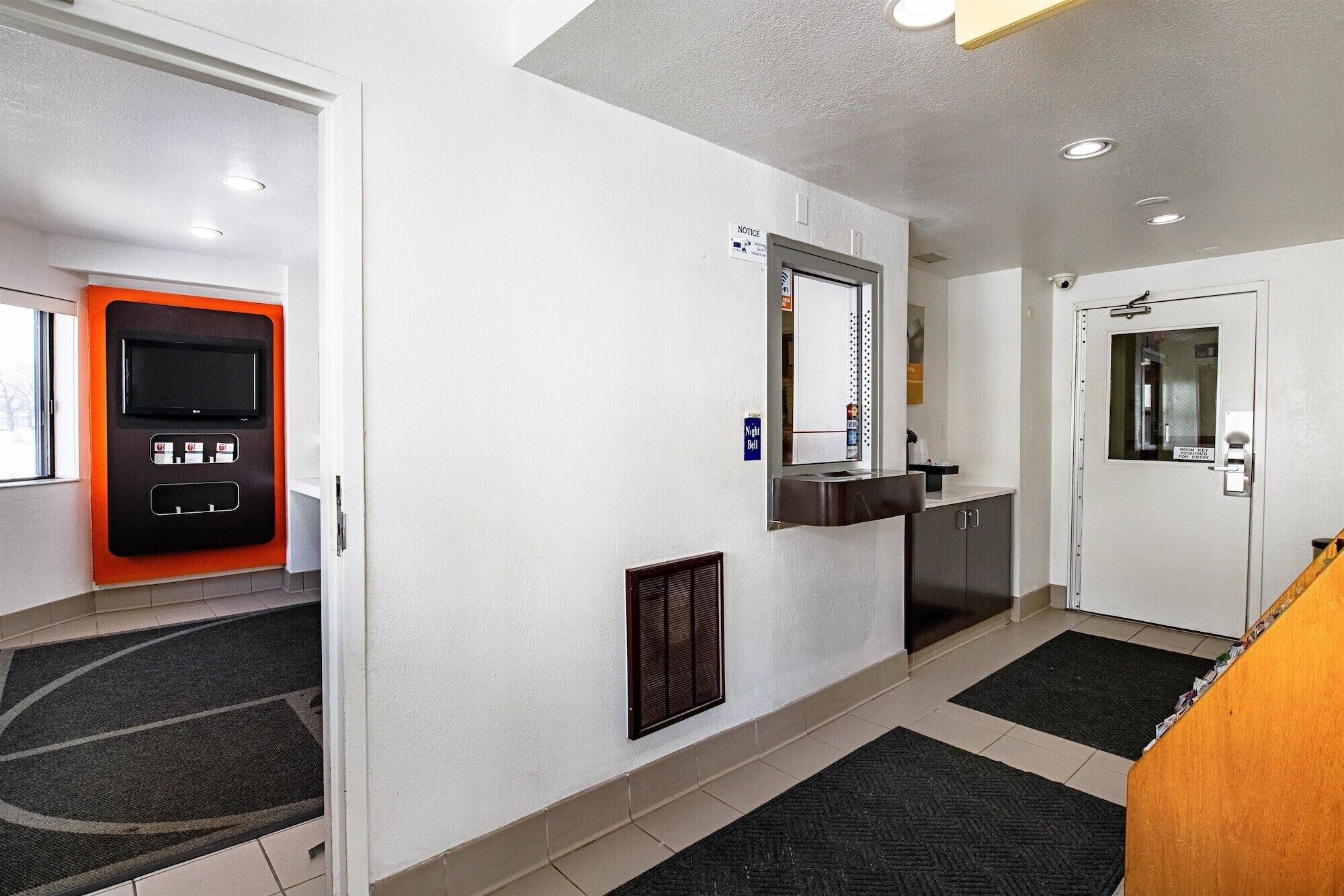 Um hall de hotel com balcão de recepção, câmera de segurança, quadro de avisos e máquina de vendas.