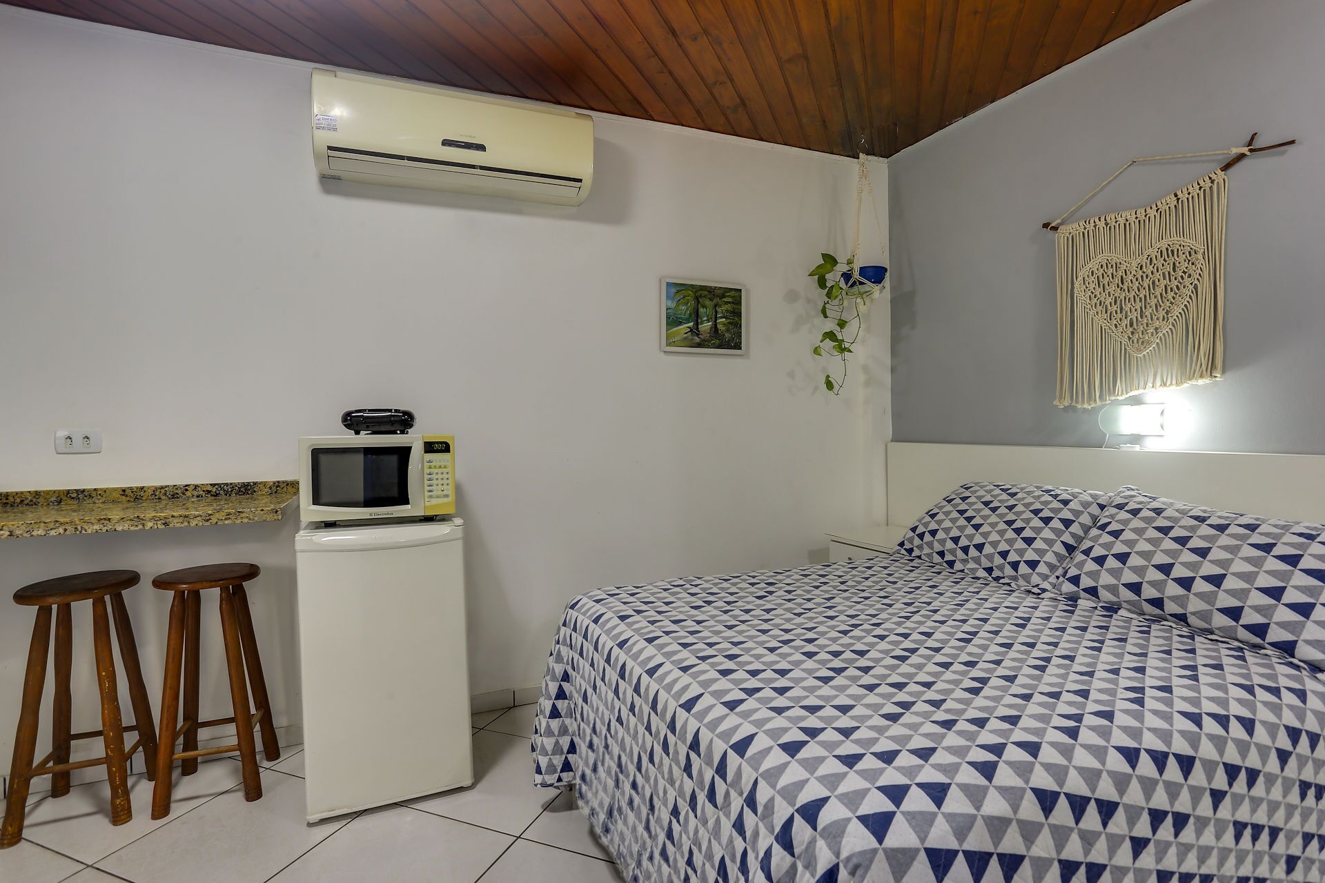 A bedroom with a bed, a microwave, and a refrigerator. There are two stools and a macrame wall hanging.