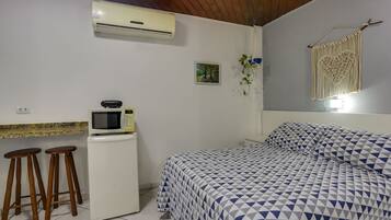 A bedroom with a bed, a microwave, and a refrigerator. There are two stools and a macrame wall hanging.