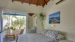 A bedroom with a bed, a nightstand, a hanging plant, and a window with a view of a cityscape. - Pousada Canário Azul (Bombinhas)